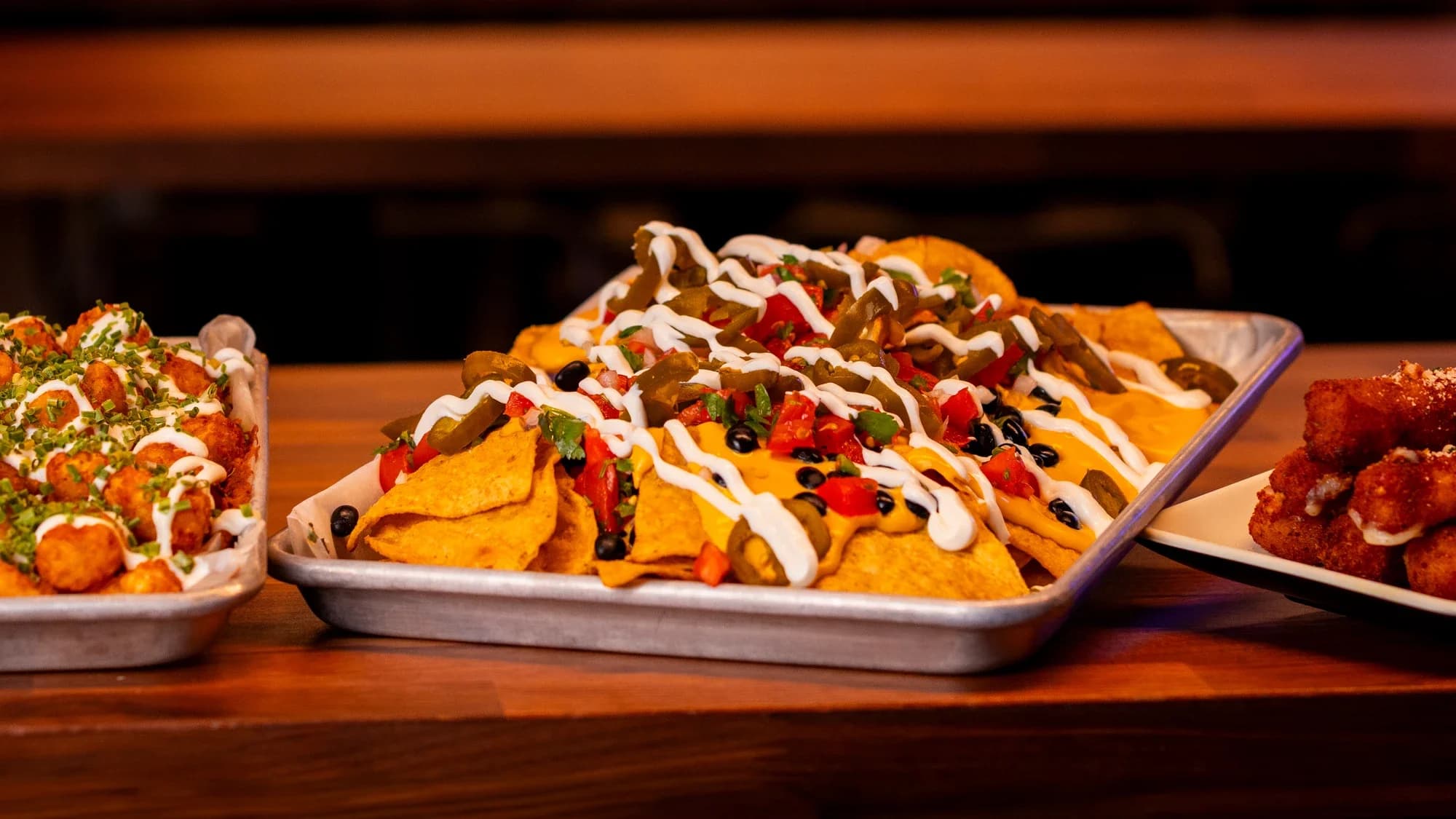 Loaded nachos, tots with sauce drizzle, and a basket of fried sticks on a wood table at Ignite Arcade.