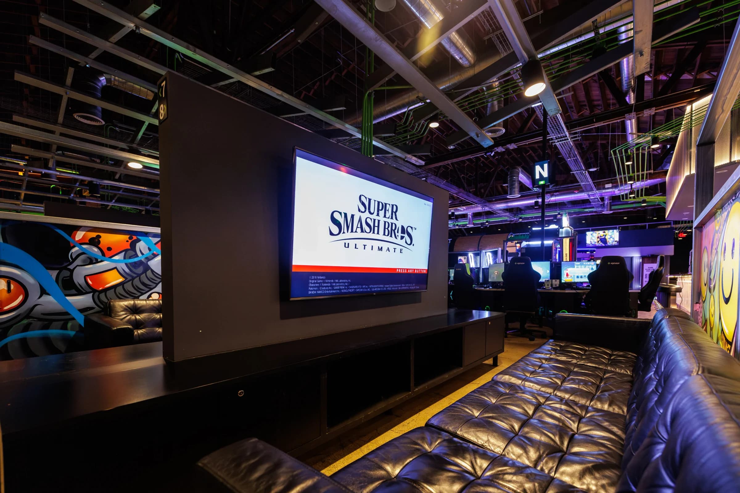 Console gaming lounge with Super Smash Bros on a big screen above a leather banquette.