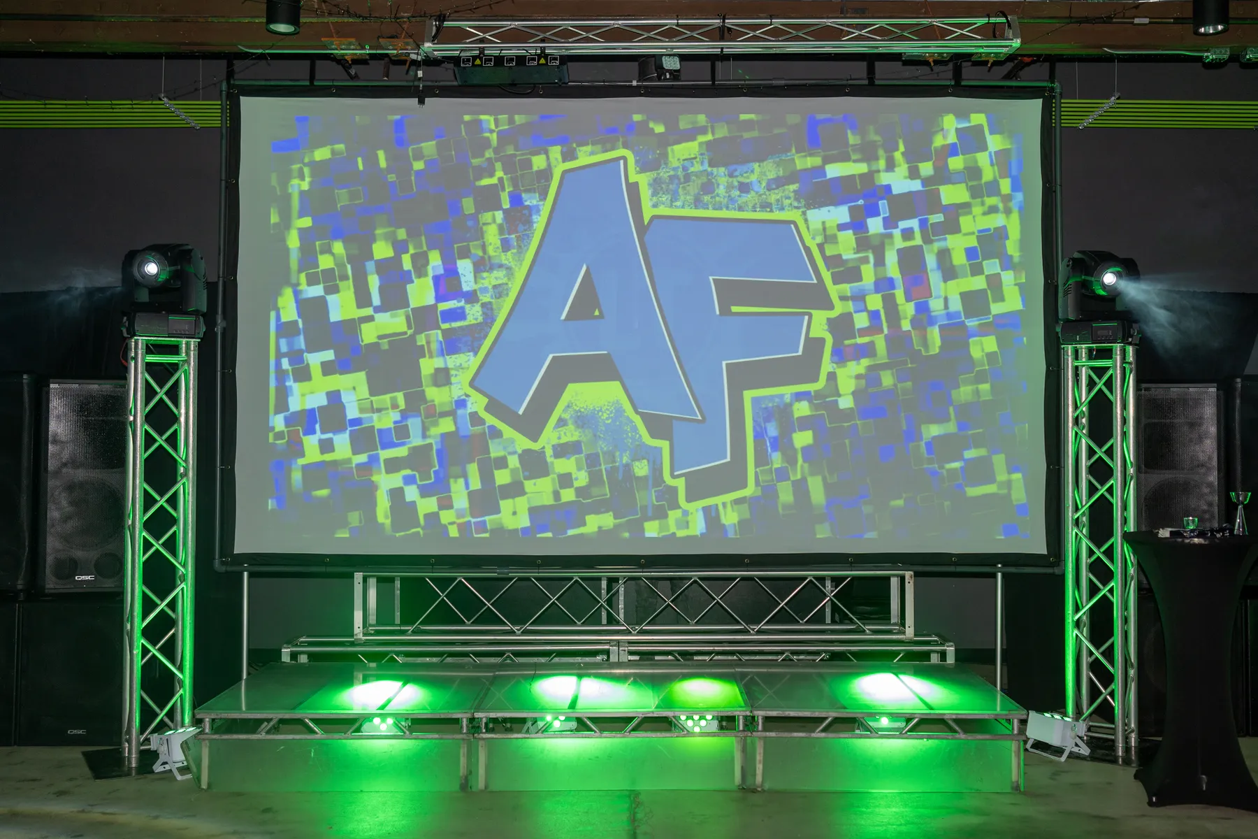 A large projector screen, stage truss, and lighting rig in the Ignite Arcade event space.