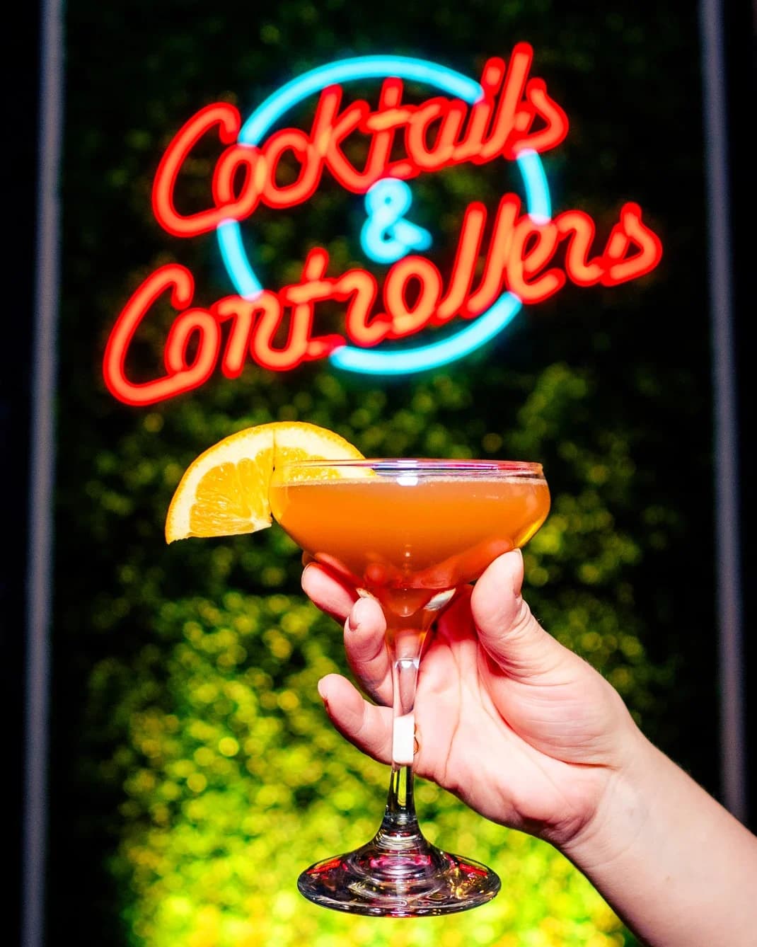 An orange cocktail held up under the Cocktails & Controllers neon sign at Ignite Arcade.